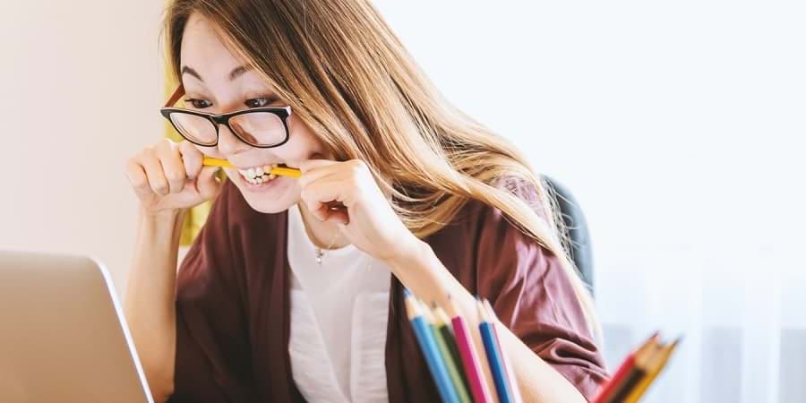 Woman wearing glasses biting a pencil, studying TCM at a laptop with colored pencils in front.