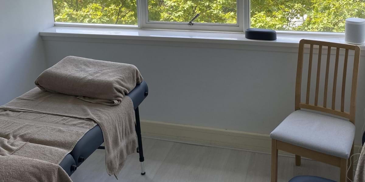 A massage table and chair for acupuncture in a bright room with speakers and a window view.
