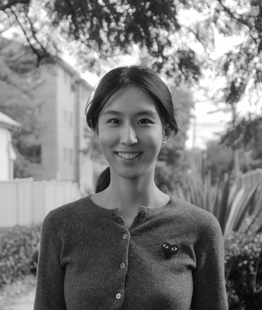 Woman smiling outdoors, with trees and buildings, evoking the calm of TCM, black and white.