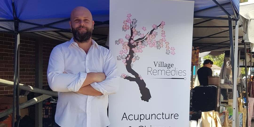 Man standing by a Village Remedies acupuncture clinic sign, promoting acupuncture and TCM.