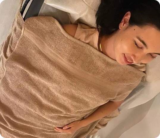 Woman lying on a spa bed, covered with a brown towel, with eyes closed and relaxed expression.