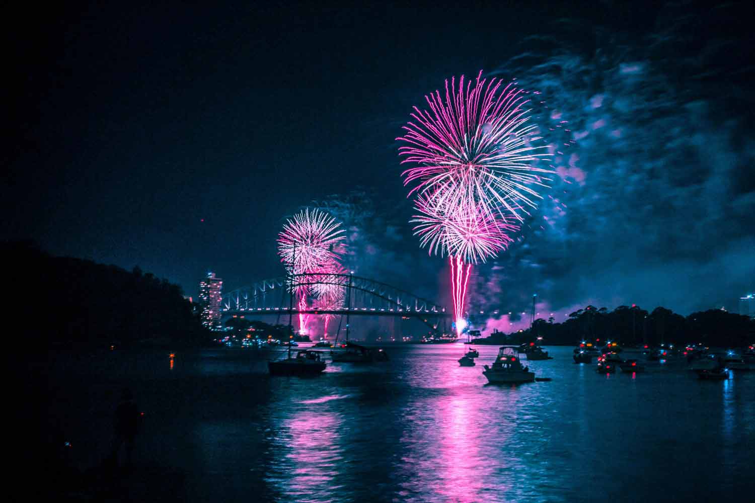 Fireworks in pink and purple light up the night sky, celebrating TCM over a river with boats below.