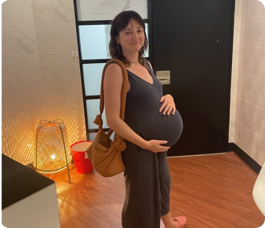 A pregnant woman stands indoors, holding her belly after receiving a pregnancy acupuncture treatment from Village Remedies