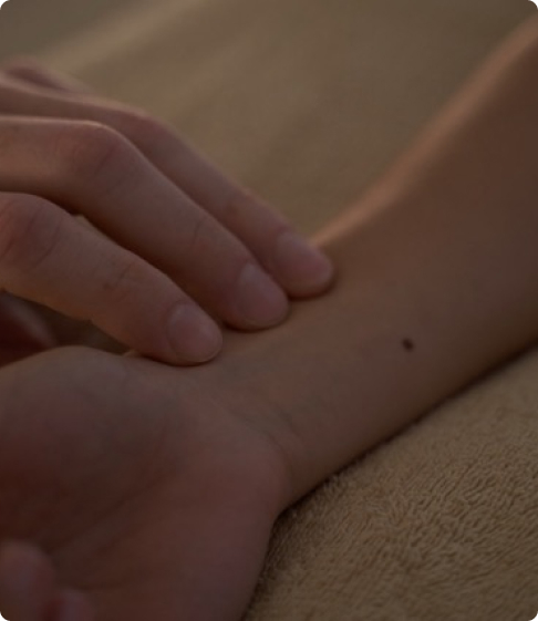 A hand touches the inside of a wrist with a visible mole on the skin.