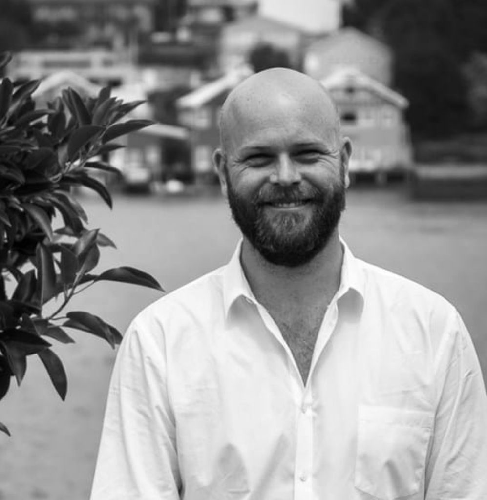A bearded man in a white shirt stands outdoors, smiling beside a tree near his acupuncture clinic.