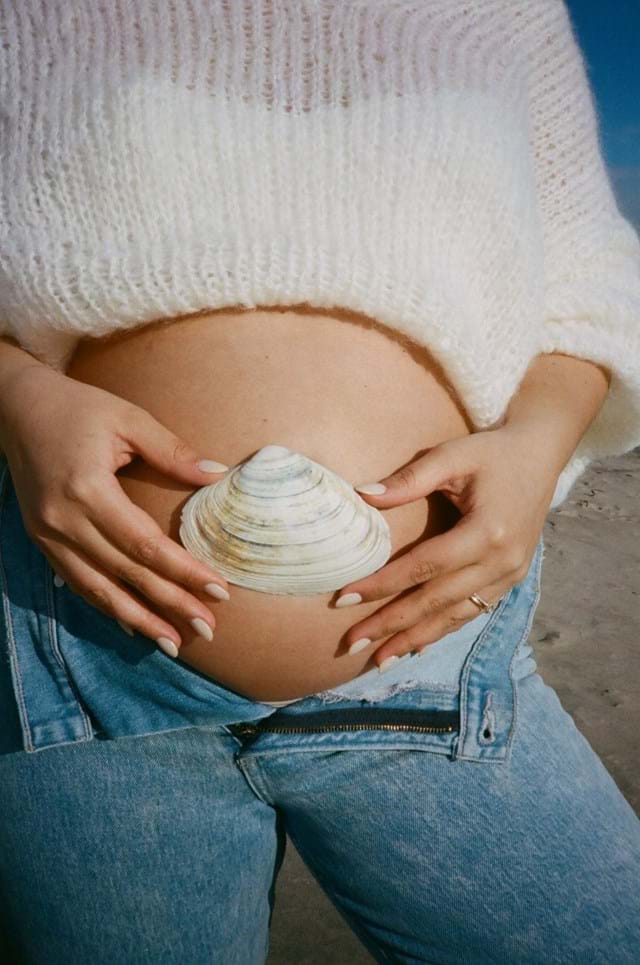 Person holding a seashell over their belly, evoking calm like tcm at the beach in jeans and a sweater.