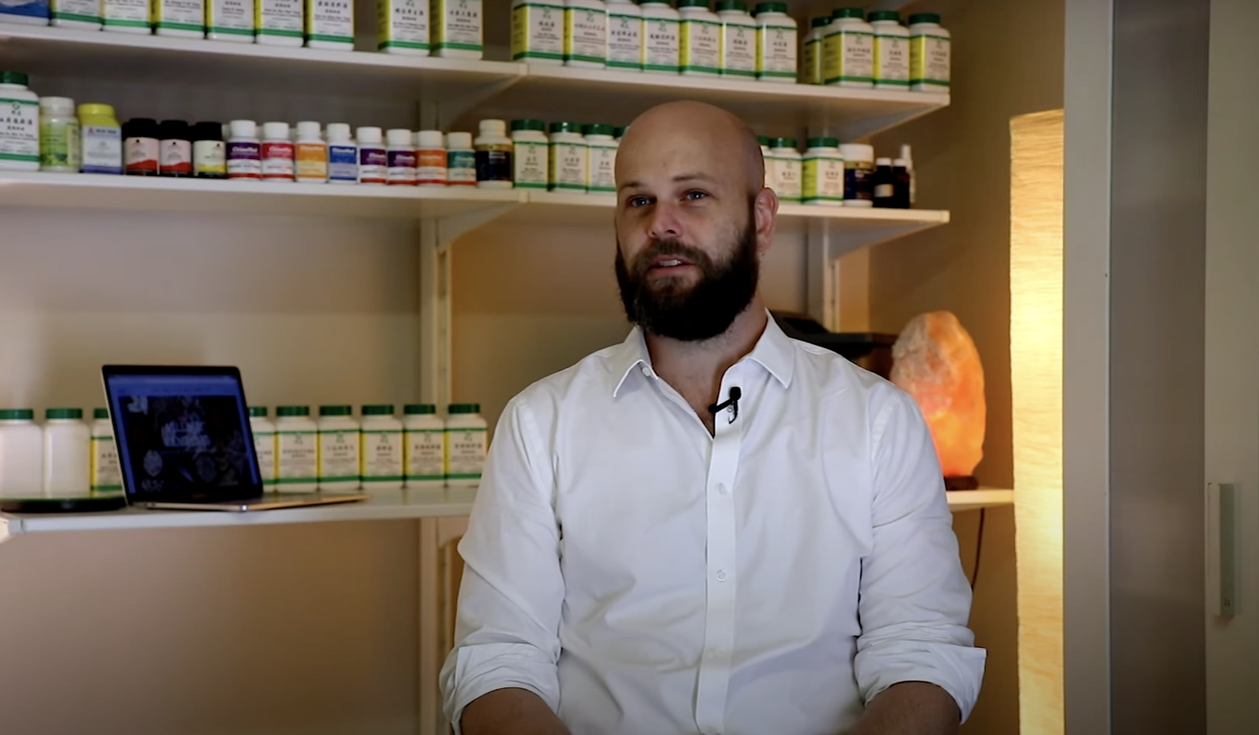 A bearded man sits in an office with shelves of Chinese herbal medicine behind him.