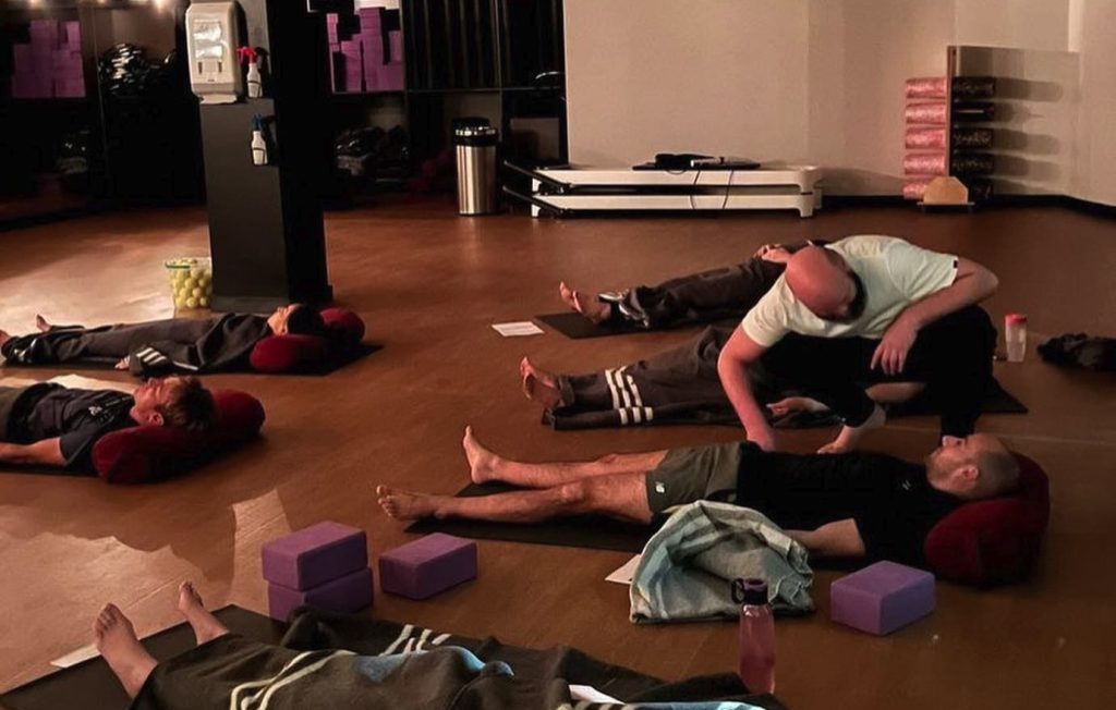 Several people lying on yoga mats in a studio; an instructor discusses acupuncture techniques.