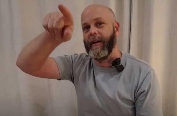 Bald man with a beard points forward, discussing acupuncture while wearing a light shirt and mic.