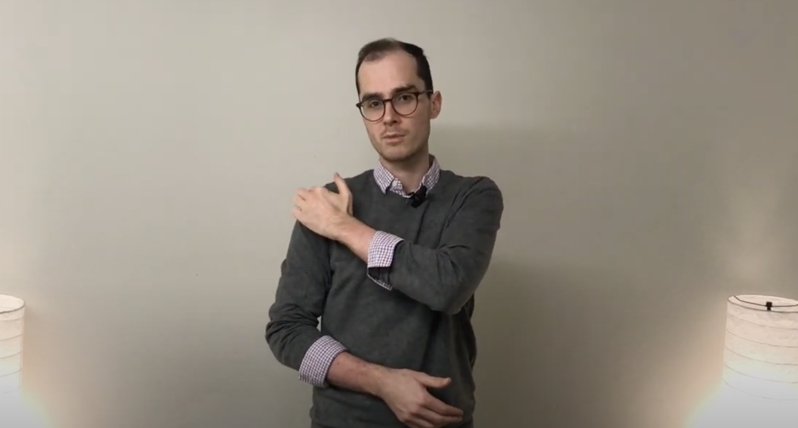 A person stands indoors, touching their left shoulder with their right hand, practicing TCM.
