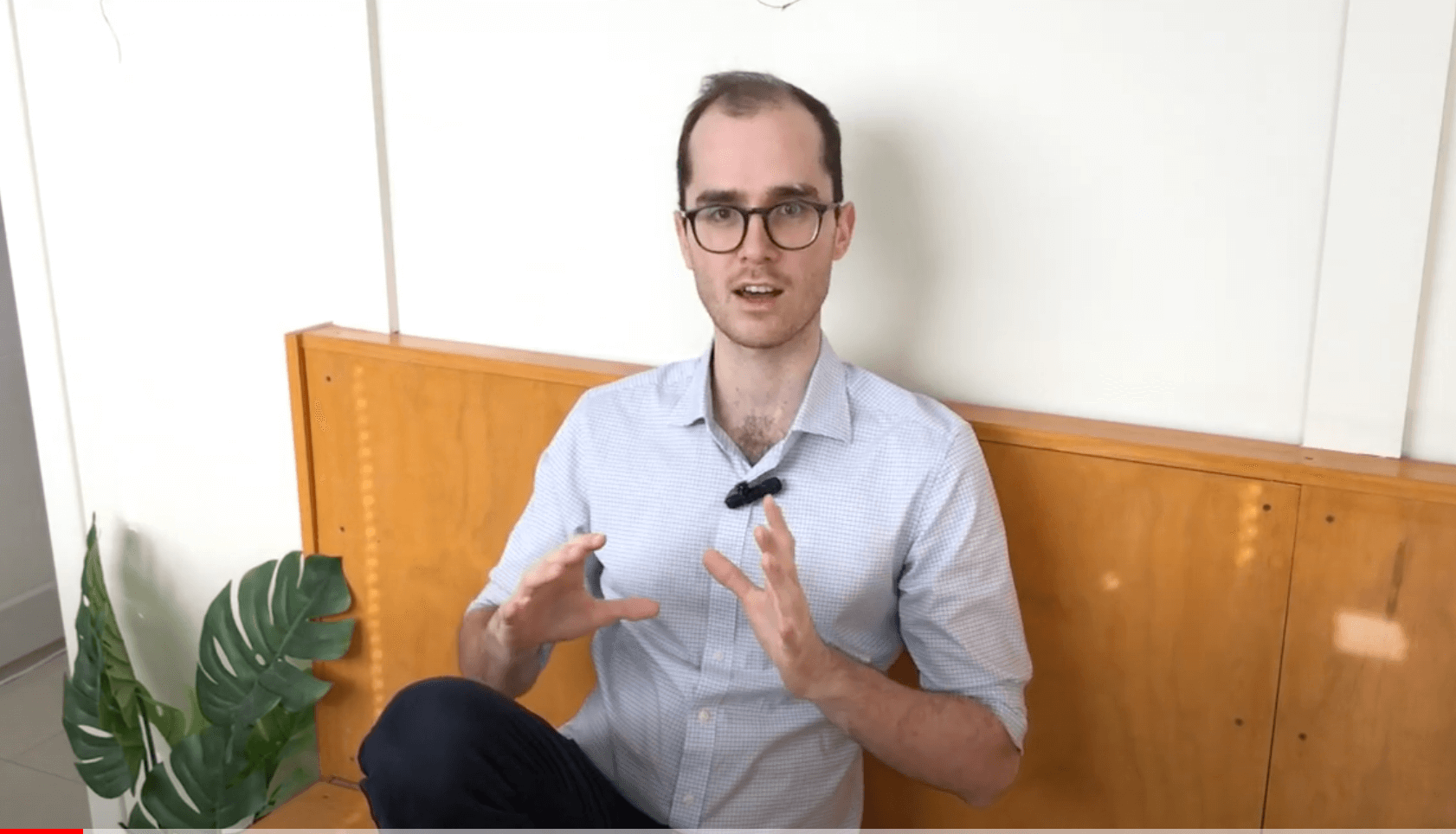 Man with glasses discusses acupuncture, gesturing on a bench next to a green plant.