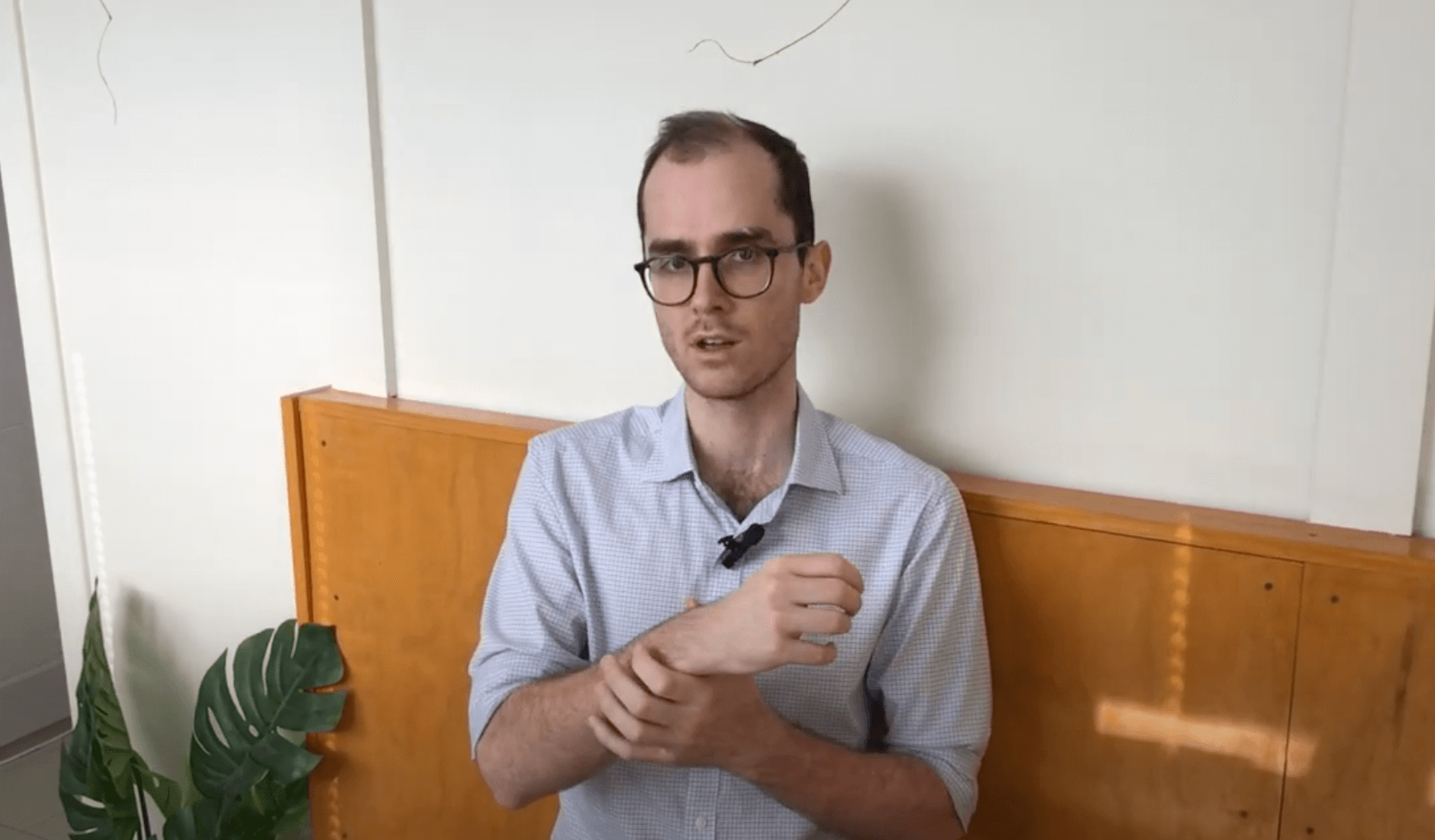 A man in glasses adjusts his sleeve, discussing acupuncture by a wooden wall.