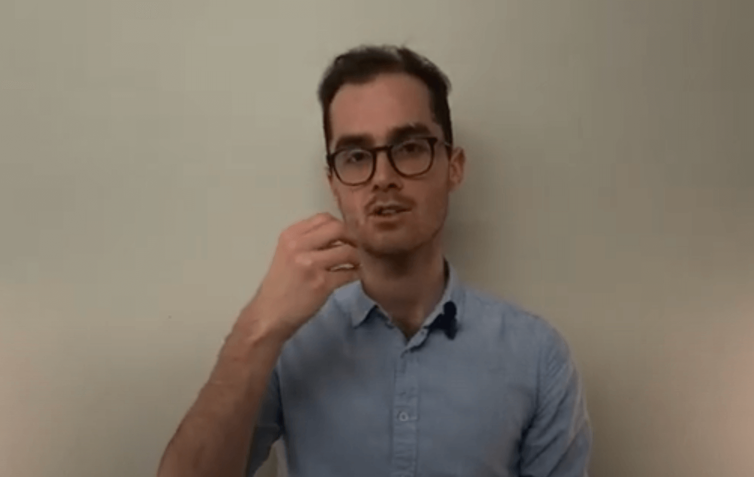 A man in glasses and a light blue shirt discusses acupuncture in front of a plain wall.