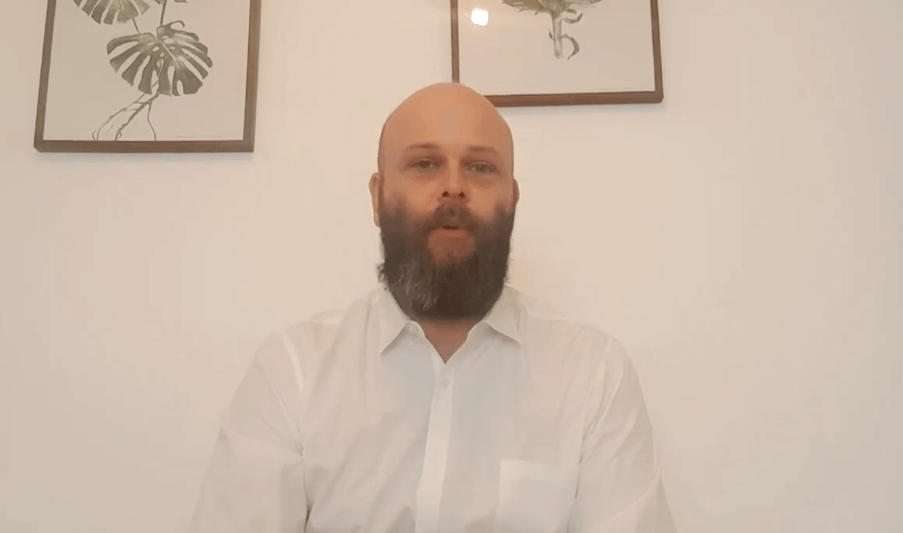 A bearded man in a white shirt sits indoors; framed TCM plant art hangs on the wall behind him.