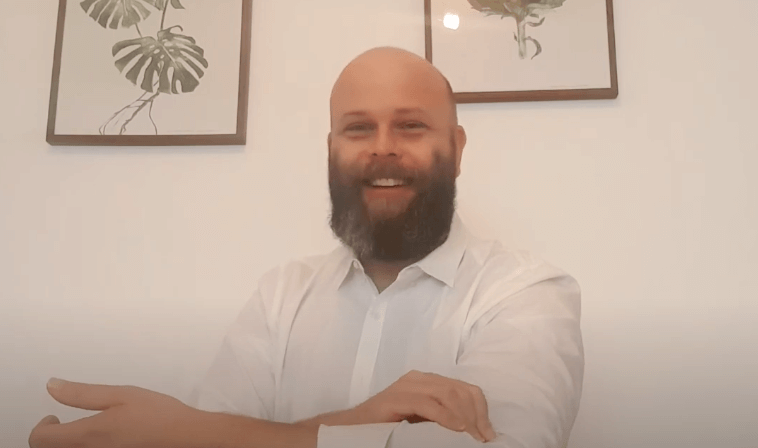 A bearded man in a white shirt smiles before framed plant artwork, evoking TCM's natural roots.