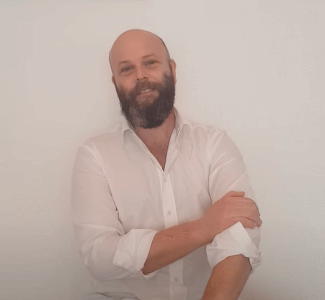 Man in white shirt smiling, rolling up his sleeve for acupuncture against a plain white background.