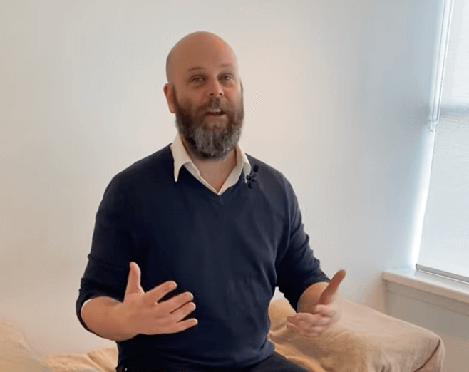A bearded man in a dark sweater gestures while discussing TCM in a bright room.