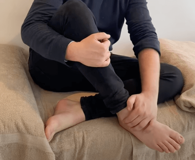 Person sitting on a bed, holding their ankle, possibly seeking TCM or acupuncture relief.