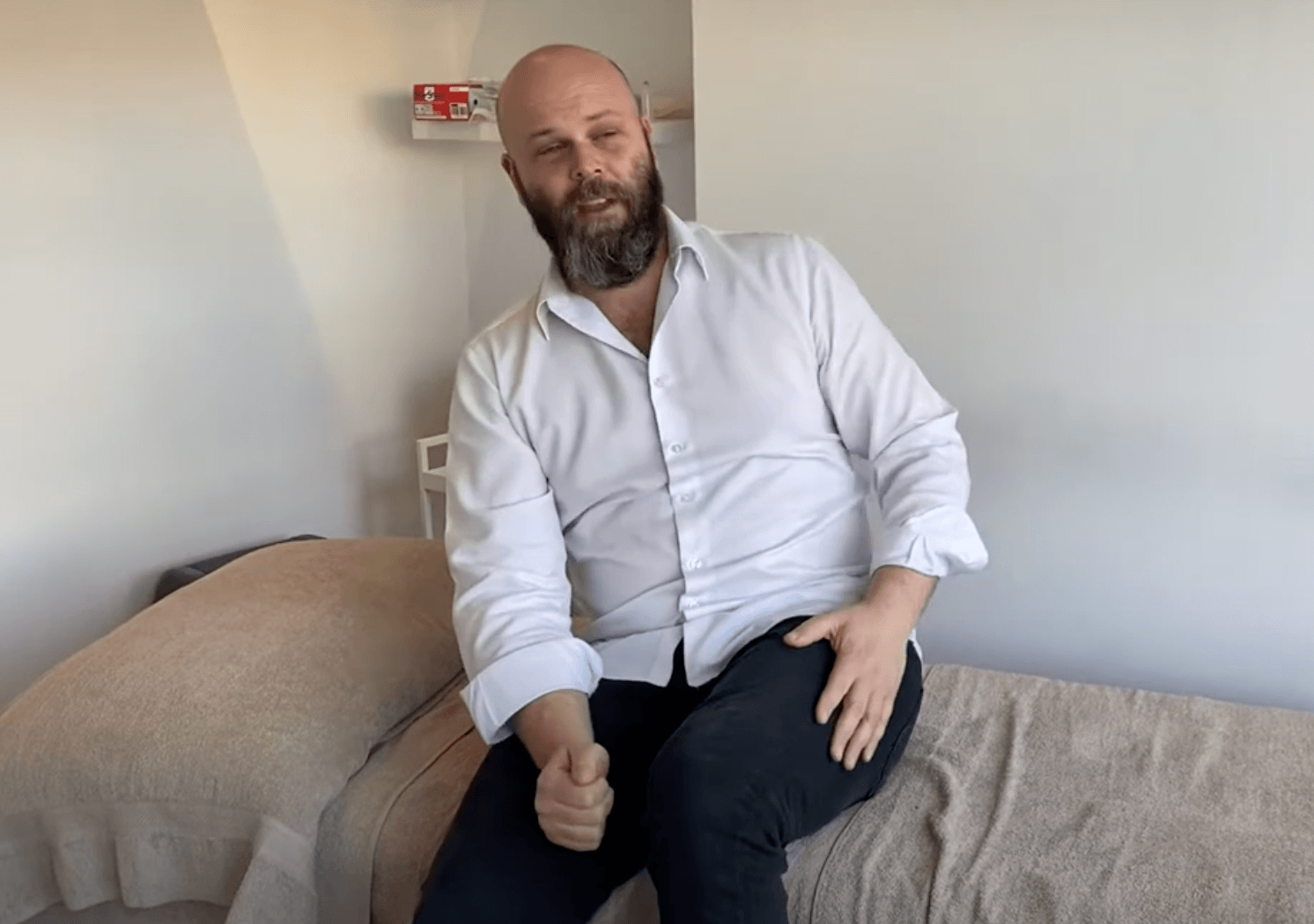 A man with a beard in a white shirt sits on a bed in a minimal room focused on TCM and wellness.