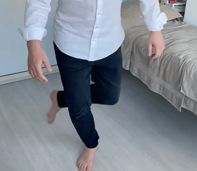 Person practicing balance on one leg indoors, inspired by TCM, near a bed in casual wear.