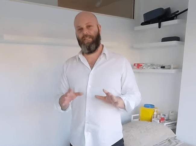 A bearded man in a white shirt stands in a medical room specializing in acupuncture supplies.