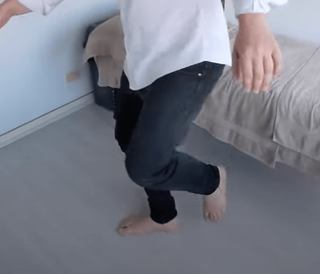 A person in jeans and a white shirt stands barefoot, practicing TCM, near a bed.