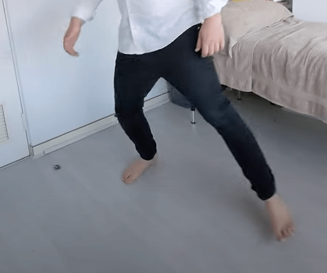 Person in a white shirt and black pants stands barefoot near a bed, evoking a TCM vibe.
