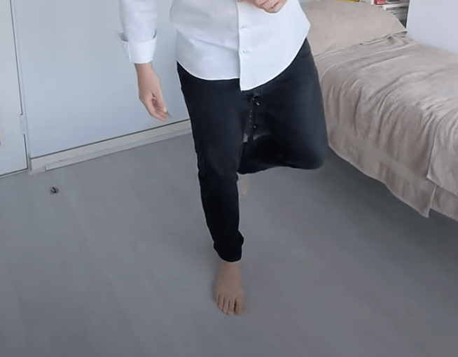 A person practicing TCM stands on one leg in a bedroom, wearing jeans and a white shirt.