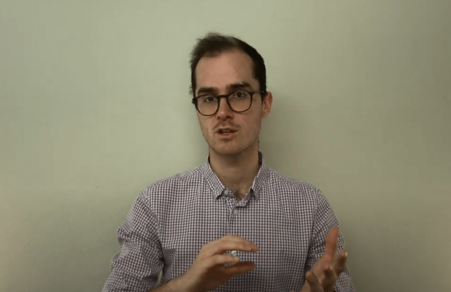 A man with glasses gestures while discussing acupuncture, standing against a light-colored wall.