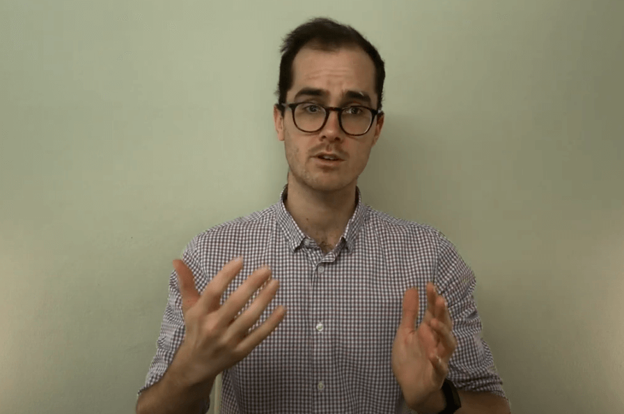 Man with glasses and checked shirt gestures, discussing acupuncture, against a light background.