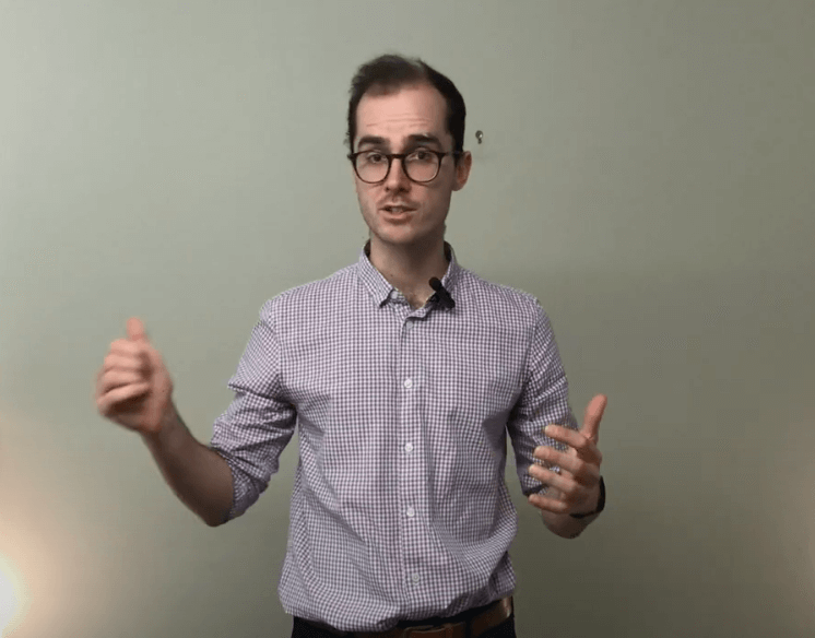 Man in glasses discusses TCM, gesturing with both hands against a plain wall.
