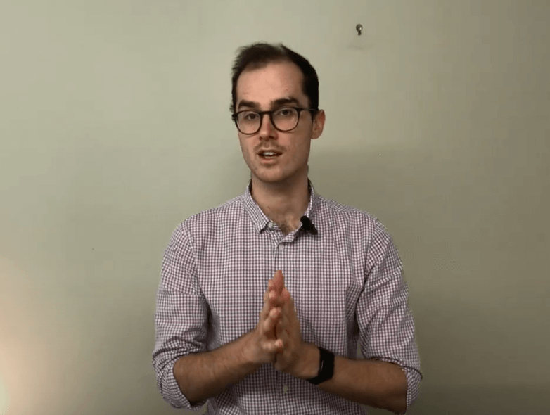 Person in glasses and checked shirt discusses acupuncture while standing against a plain wall.