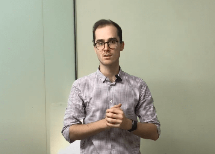 A man with glasses stands indoors, clasping his hands in a TCM clinic and facing the camera.