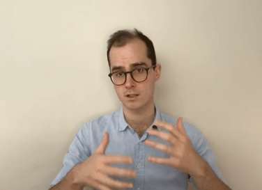 A man in glasses gestures while discussing acupuncture in front of a plain wall.