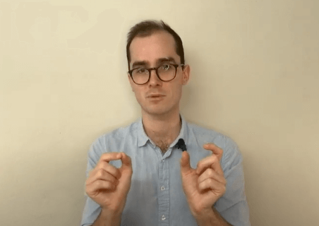 A person wearing glasses gestures with both hands, explaining acupuncture, against a plain wall.