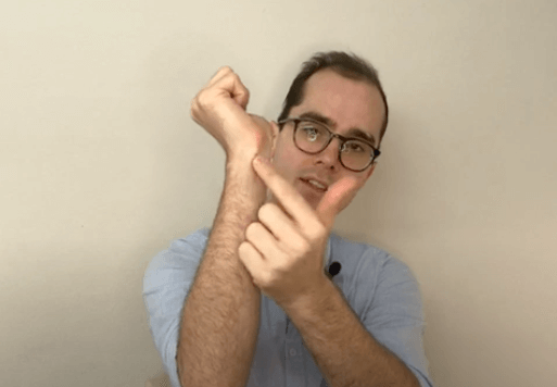A man points to his wrist, demonstrating an acupuncture point, in front of a plain background.