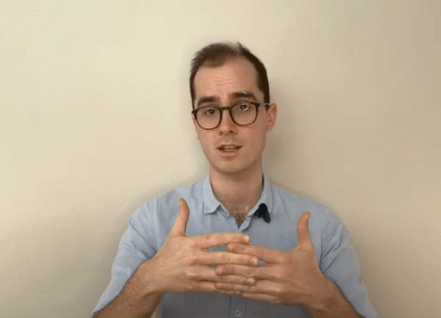 Man with glasses in a blue shirt discusses TCM while gesturing against a plain background.
