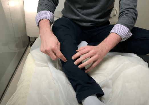 Person points to their shin, highlighting an acupuncture point while sitting cross-legged on a bed.