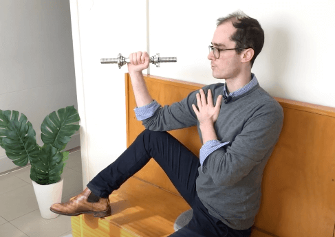A man in glasses holds a barbell, touching his chest on a wooden bench after acupuncture.