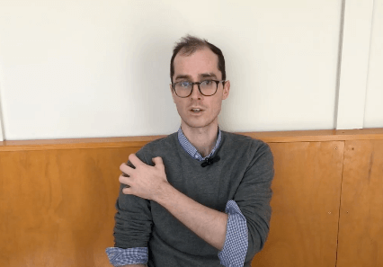 Man in glasses and gray sweater sits by a wood-paneled wall, seeking relief with acupuncture.