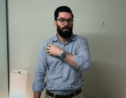Man with glasses and beard touches his shoulder, considering acupuncture by a lamp and blank wall.