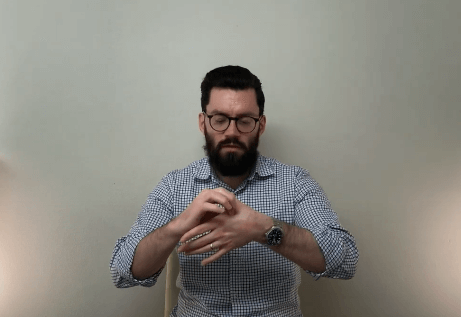 A man with glasses and a beard signs in front of a plain wall about acupuncture.