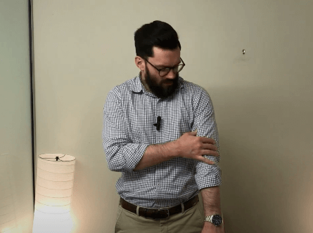 Man with rolled-up sleeves adjusts his shirt near a floor lamp in a TCM-inspired office.