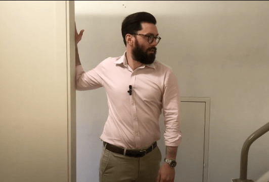 Man in glasses and pink shirt stands near a doorway, next to shelves of Chinese herbal medicine.