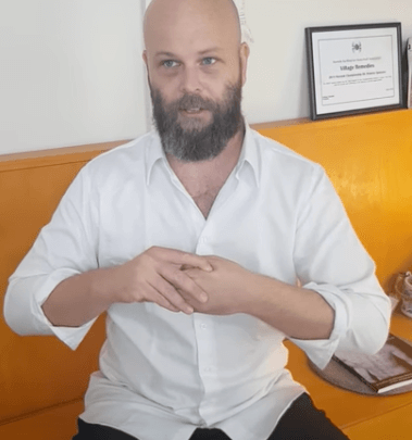 Man with a beard sits on a yellow bench, wearing a white shirt, discussing acupuncture.