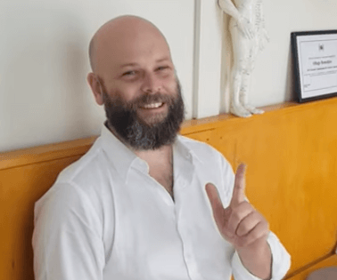Man with a beard in a white shirt smiles and gestures, sharing about TCM while seated indoors.