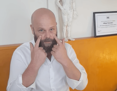 Man in white shirt points to his eyes, hinting at tcm or acupuncture in a yellow-walled room.