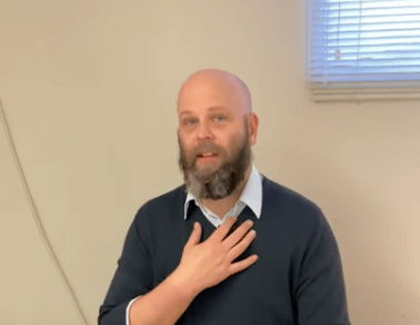 A man with a beard touches his chest, reflecting on tcm in a room with window and blinds.