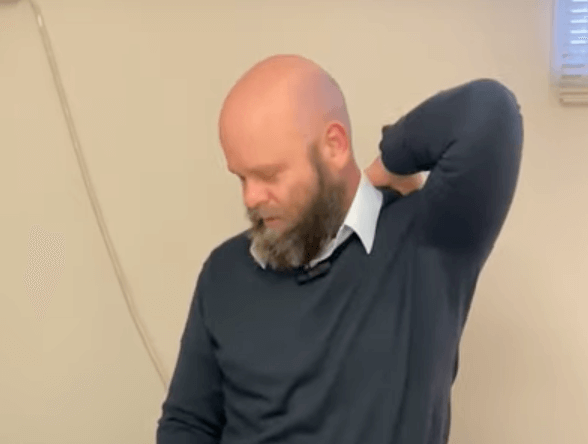 Man with a beard touches his neck, appearing pensive in an indoor tcm setting.