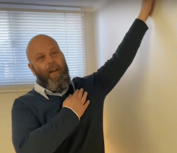 A man indoors touches his chest, reaching up as if considering acupuncture or tcm practices.